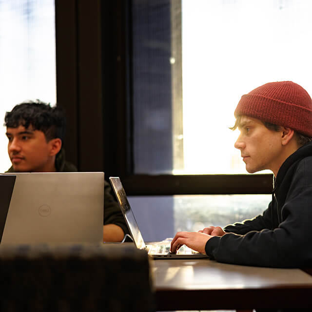 Two students sit at a table studying on their laptops.