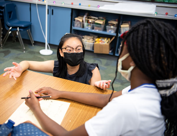 A student teacher kneels to help a student at her desk.