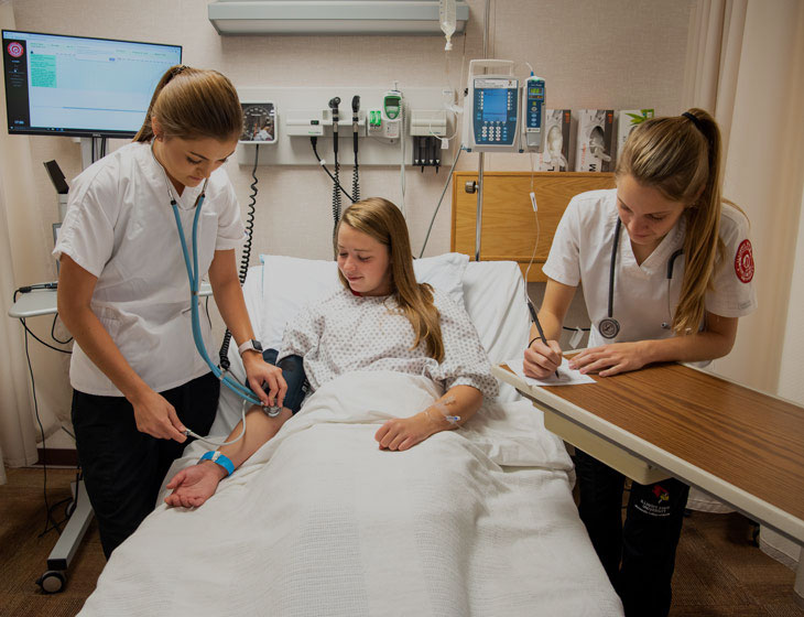 Two nursing students practice examining their peer while she lays in a hospital bed.
