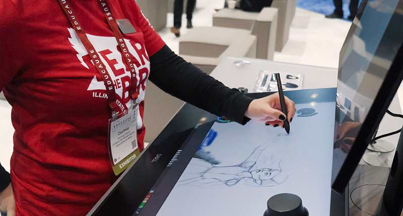 Female sketching a person using an interactive pen on a large tablet at a convention.