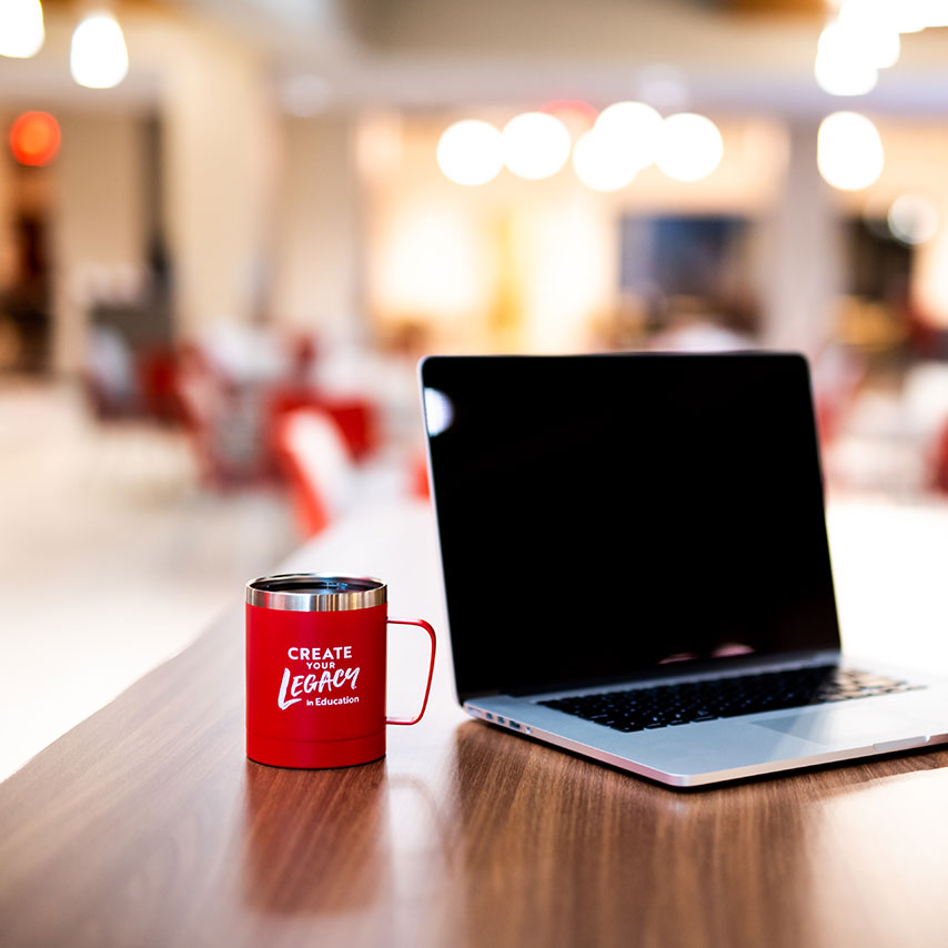 A laptop and a red mug on a wooden table in a softly lit room.