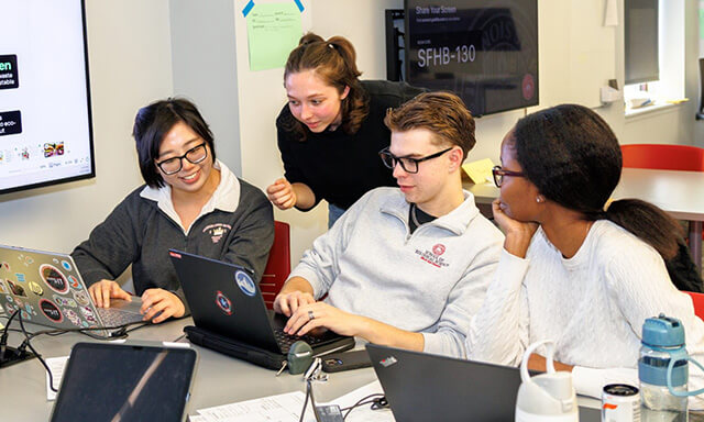 Four individuals collaborating around laptops in a classroom setting.