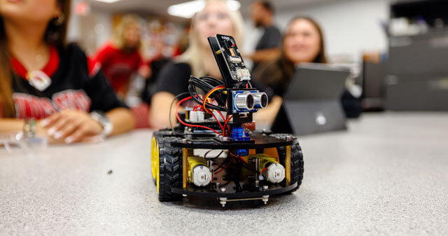 A small robotic vehicle with electronic components on a table, with people in the background.