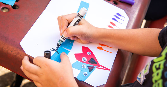 Close-up of a child’s hands creating a colorful paper airplane craft with markers and decorations.