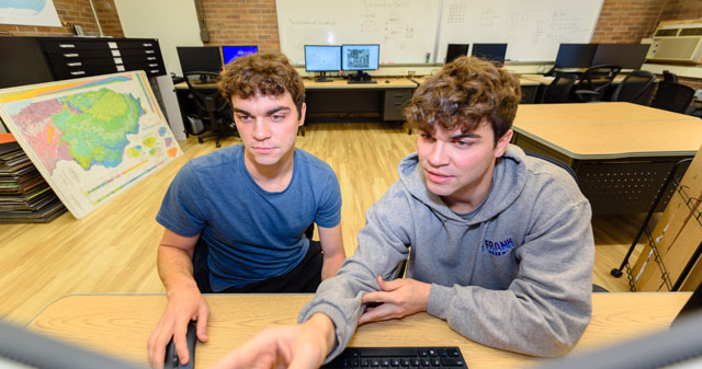 Two young men looking at a computer screen in a classroom with multiple monitors and a map in the background.