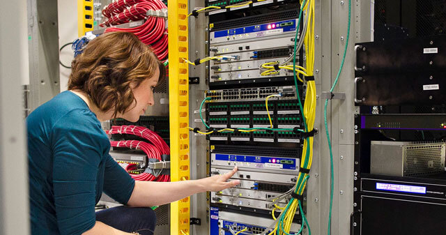 A woman in a server room interacts with electronic equipment in a server rack, surrounded by colorful cables.