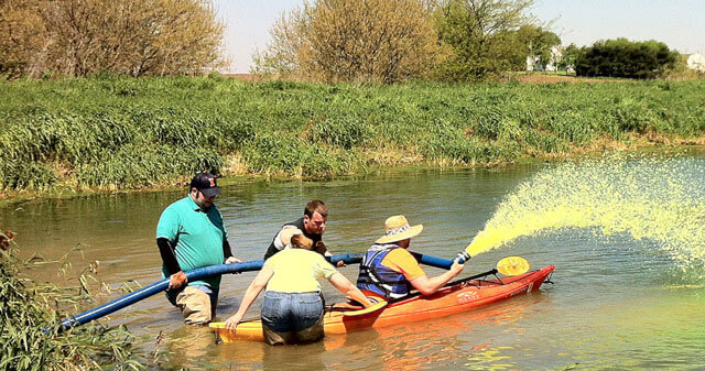 A group of people assisting a man spraying yellow liquid from a kayak on a pond.