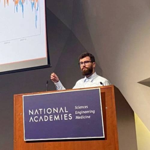 Joseph Wittrock stands behind a lectern while presenting. A screen displaying a line graph is over his shoulder.