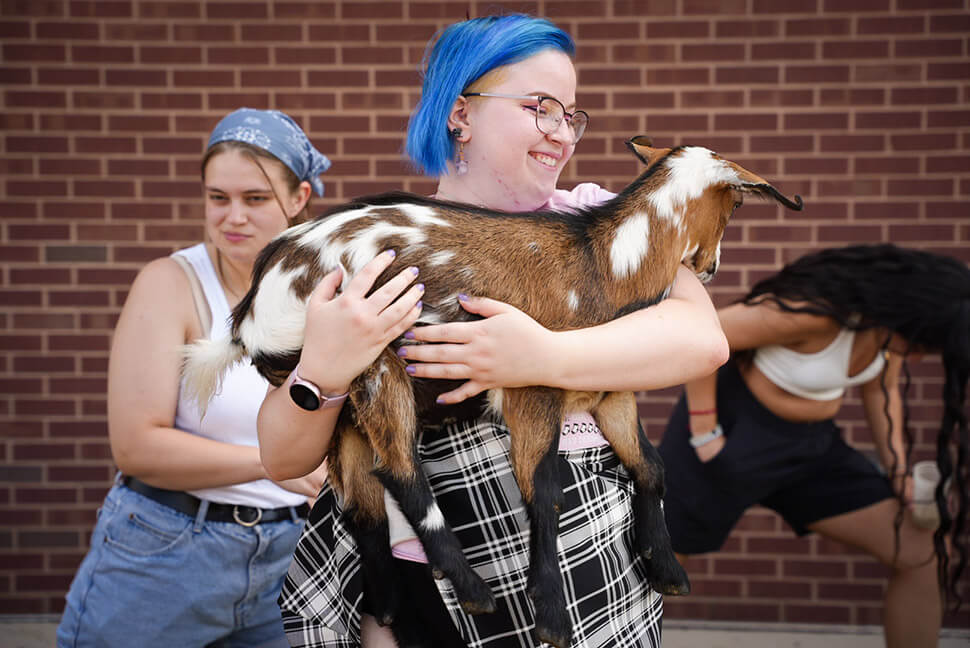 Person with blue hair holding a goat at Goat Yoga in the Brickyard Plaza.